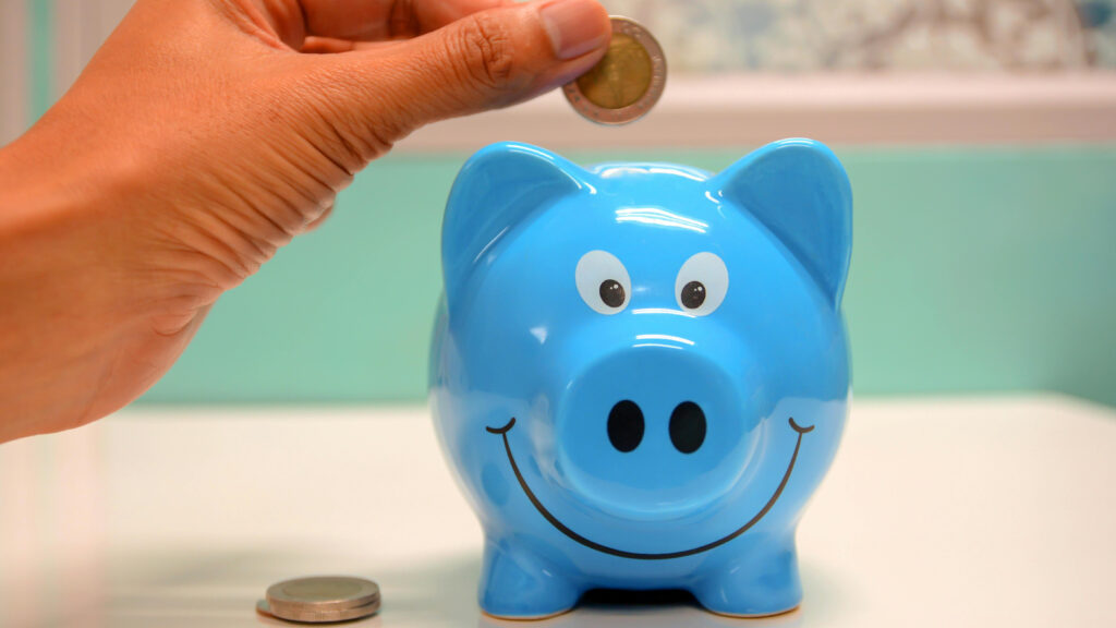 A hand placing a coin into a blue piggy bank with a smiling face.