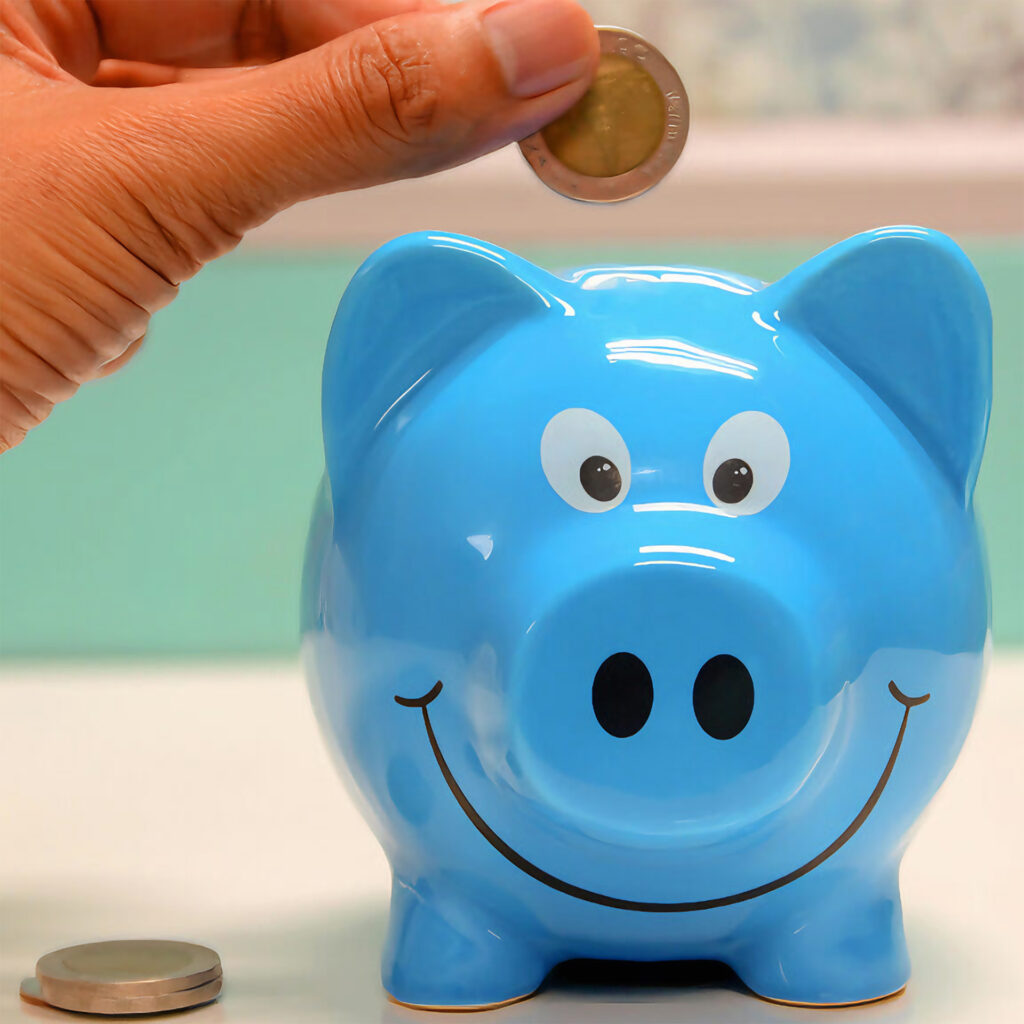 Hand placing a coin into a blue piggy bank, symbolizing saving money.